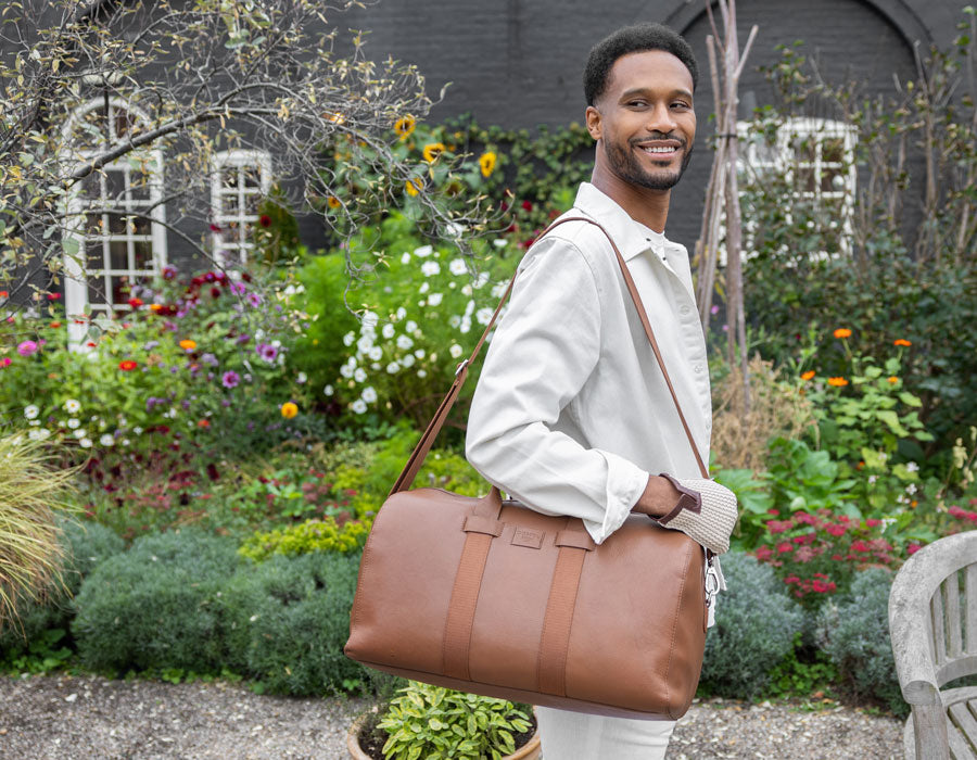 Man holding a caramel leather weekend bag in a garden setting