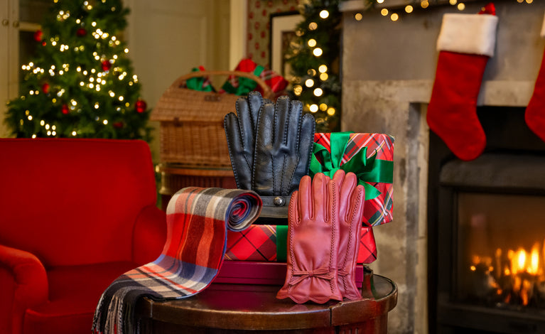 A cosy holiday scene with a round table displaying a pair of men's black leather gloves, a pair of women's red leather gloves, a cashmere plaid scarf, and a wrapped gift, set in front of a fireplace with red stockings and a Christmas tree.