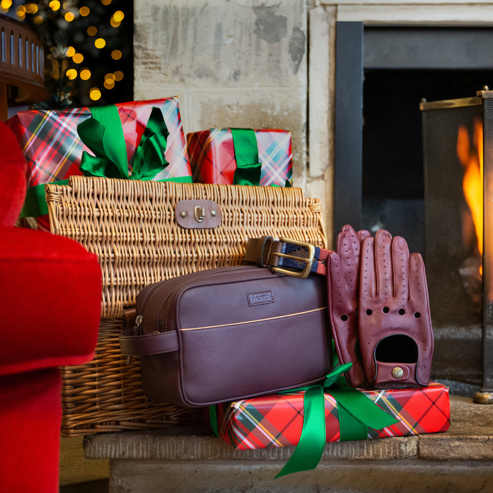 Men's brown leather driving gloves, a men's leather wash bag and polo belt with a wrapped gift present and wicker hamper in front of a roaring fireplace