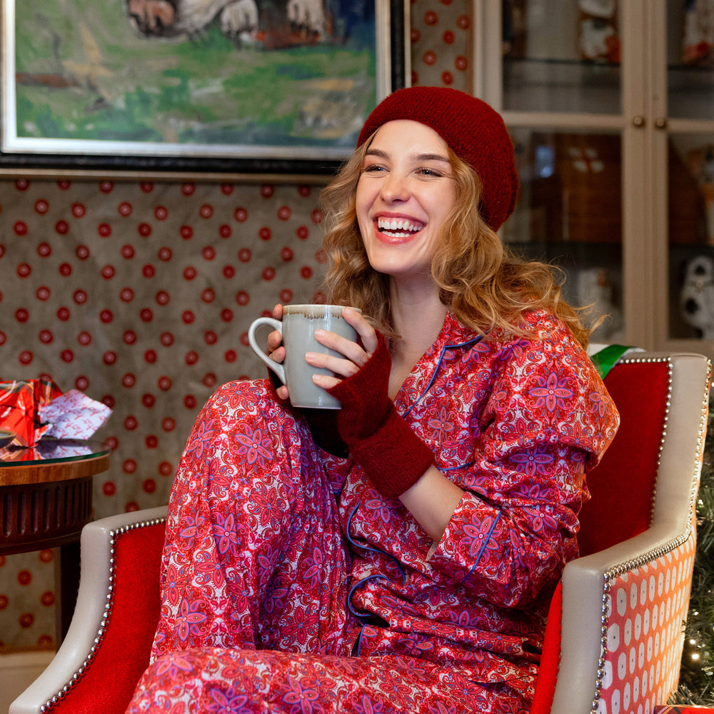 A woman wearing a burgundy knitted beanie hat and matching wrist warmers is sat on a red chair in her pajamas holding a grey mug on Christmas morning.