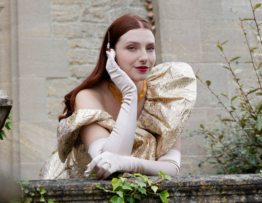 Woman in a gold dress with white gloves standing outdoors against a stone wall.