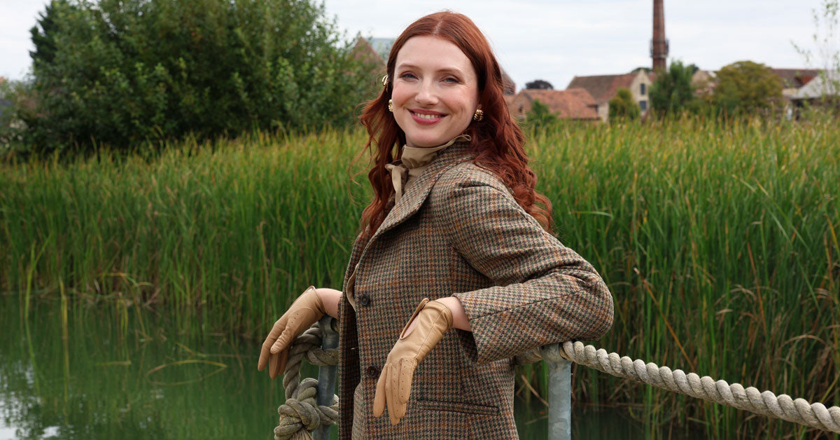 Woman in a checkered coat standing by a lake with greenery in the background wearing driving gloves in toffee